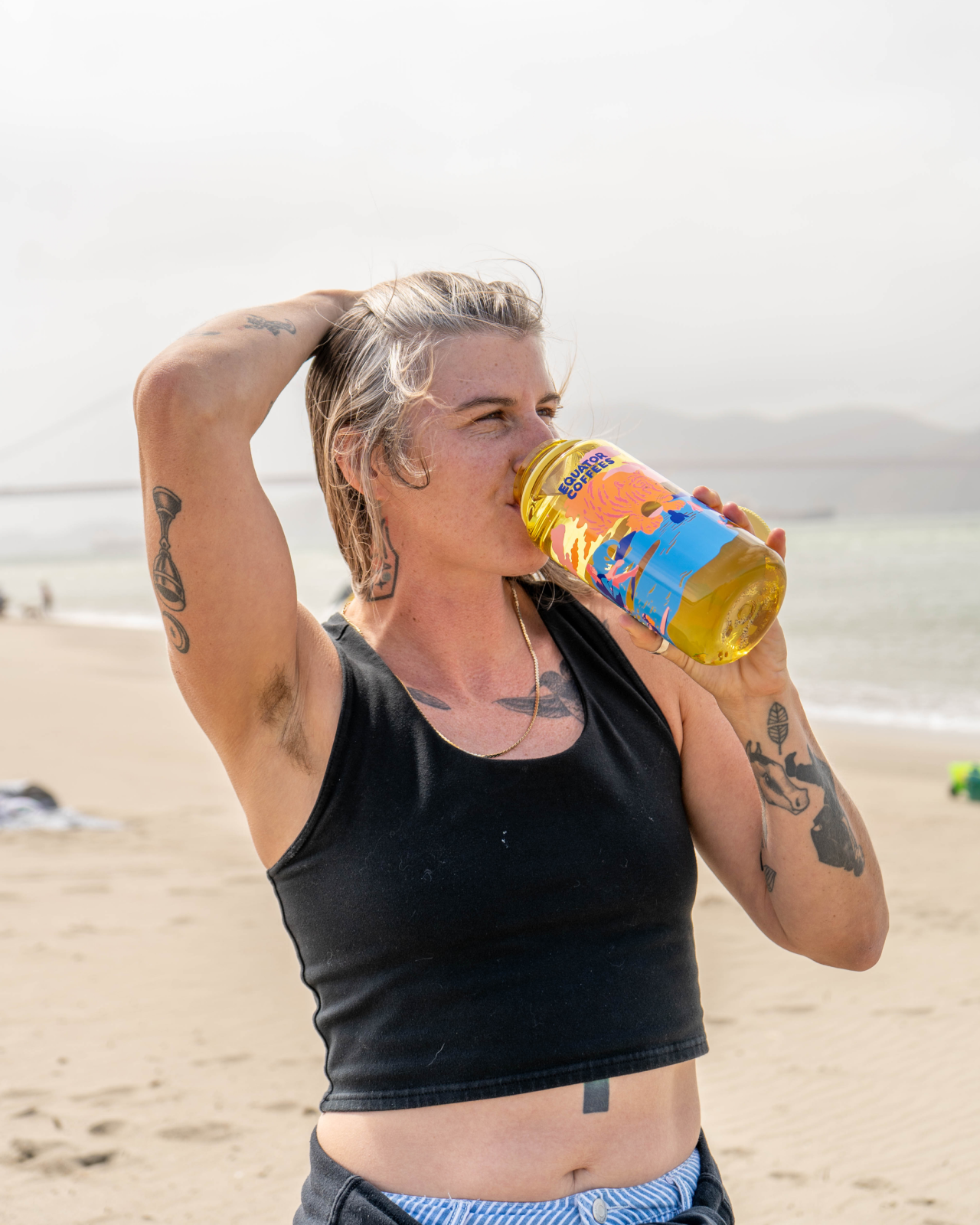 A woman on a beach drinking from a yellow and blue Golden Hour Nalgene bottle | Equator Coffees